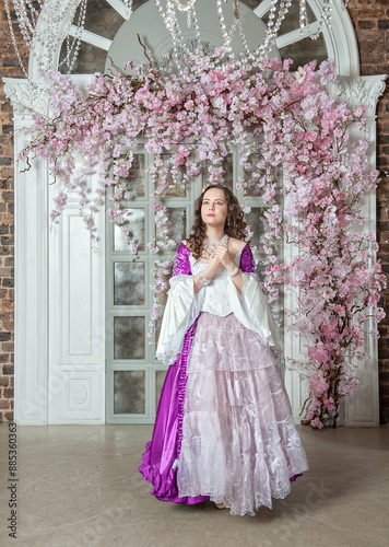 Beautiful woman in fantasy white and purple rococo style medieval dress standing near wall with pink flowers