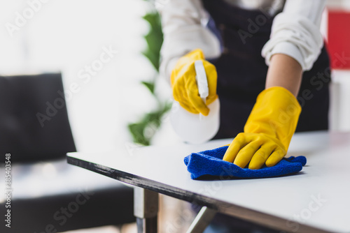 A young woman wearing an apron is cleaning a table in her home office using disinfectant and a wipe.