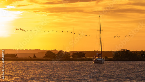 a boat with its mast down sitting in the water during sunset in the marsh with birds flying overhead