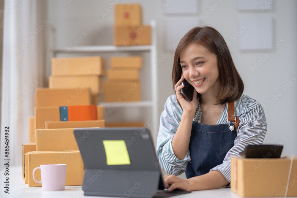 Smiling young woman running a small business from home, talking on the phone, using a laptop, and surrounded by cardboard boxes. Smiling young woman running a small business from home, talking on the phone, using a laptop, and surrounded by cardboard boxes.