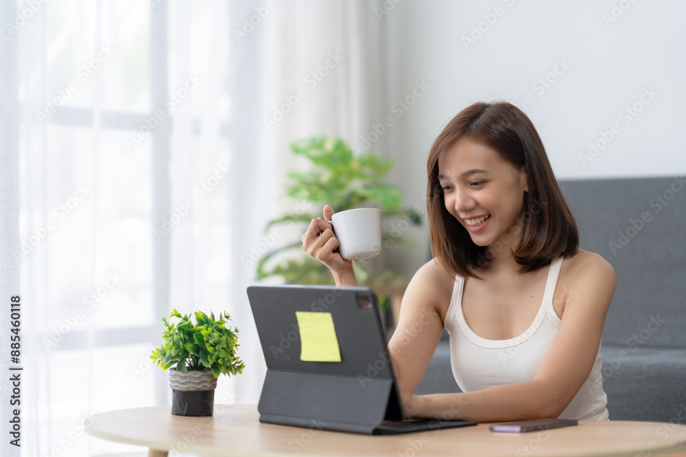 Smiling woman holding coffee cup while working on laptop at home. Cozy remote work environment with green plants and bright natural light. Smiling woman holding coffee cup while working on laptop at home. Cozy remote work environment with green plants and bright natural light.