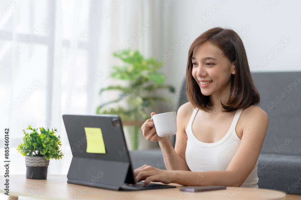 Smiling woman working on laptop at home while drinking coffee, sitting on couch with a plant and phone on the table. Smiling woman working on laptop at home while drinking coffee, sitting on couch with a plant and phone on the table.
