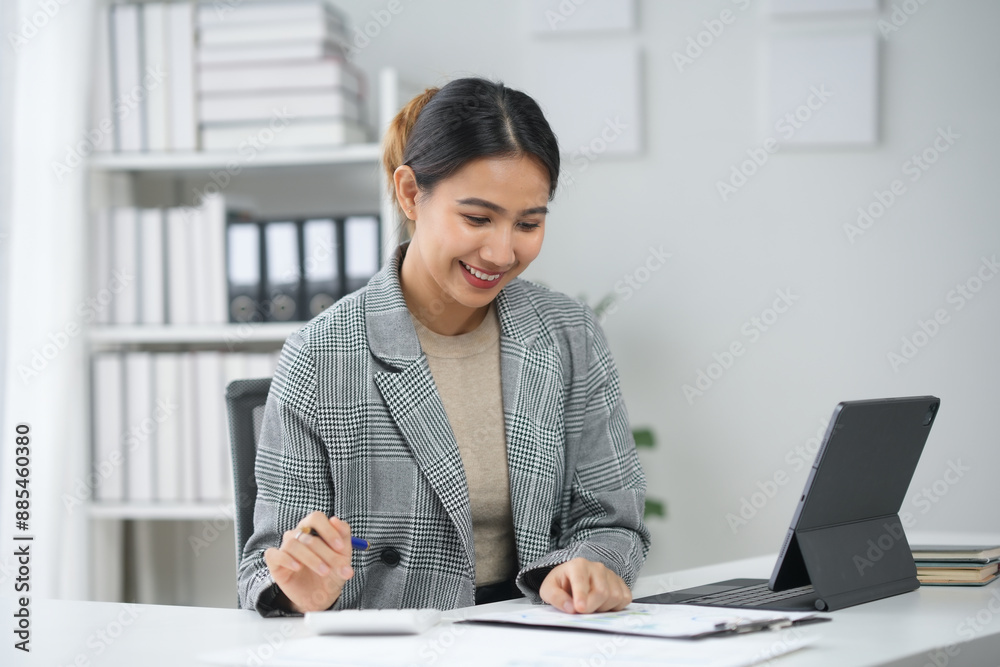 Businesswoman working on a desk with a laptop, reviewing documents, smiling, in an office setting. Businesswoman working on a desk with a laptop, reviewing documents, smiling, in an office setting.