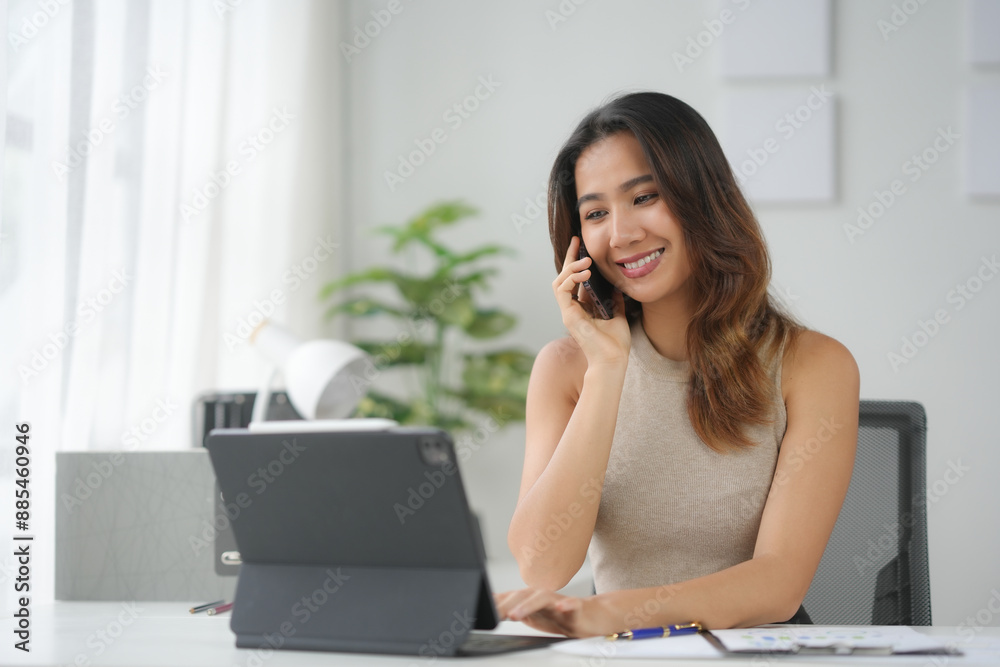 Smiling woman working in a modern office setting, talking on the phone, using a laptop, surrounded by paperwork and plants. Smiling woman working in a modern office setting, talking on the phone, using a laptop, surrounded by paperwork and plants.