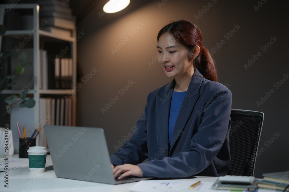 Businesswoman working late at night in office, typing on laptop. Professional and focused. Dimly lit workspace with documents and coffee.