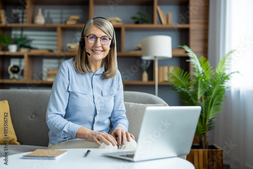 Smiling senior woman wearing headphones with microphone, working remotely from home laptop. Confident professional communicating online, participating virtual meeting. Cozy home office environment.