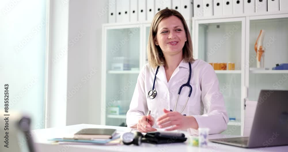 Pleased doctor writes notes in patient history distracting on patient entering office. Positive woman in medical uniform sits at table preparing for appointment Pleased doctor writes notes in patient history distracting on patient entering office. Positive woman in medical uniform sits at table preparing for appointment
