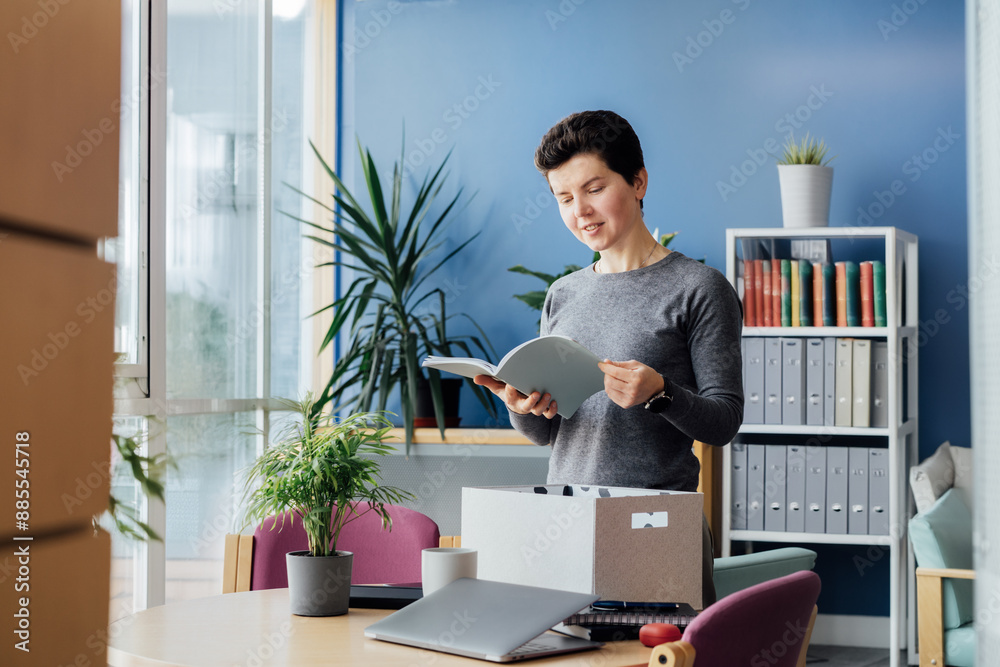 Motivated newcomer middle aged neutral gender female standing in office unpacking box with personal belongings, checking project plan at workplace having first working day concept. New Carrier start Motivated newcomer middle aged neutral gender female standing in office unpacking box with personal belongings, checking project plan at workplace having first working day concept. New Carrier start