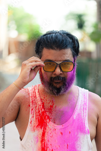 Close-up of a bearded man wearing sunglasses, covered in Holi colors, looking at the camera during the festival of Holi.