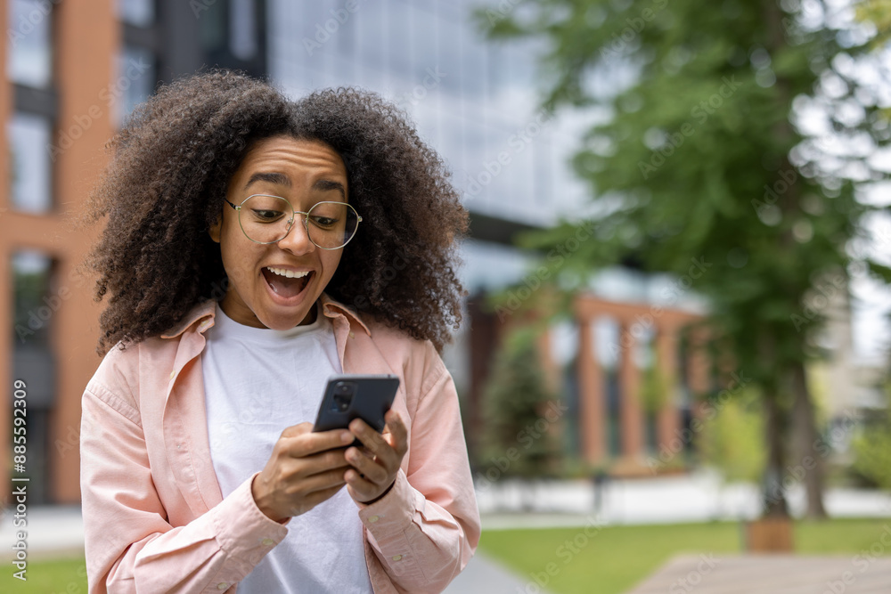 Joyful woman student with curly hair holding smartphone outdoors on campus. Young woman using mobile phone, looking pleasantly surprised and happy. Urban outdoor setting. Joyful woman student with curly hair holding smartphone outdoors on campus. Young woman using mobile phone, looking pleasantly surprised and happy. Urban outdoor setting.
