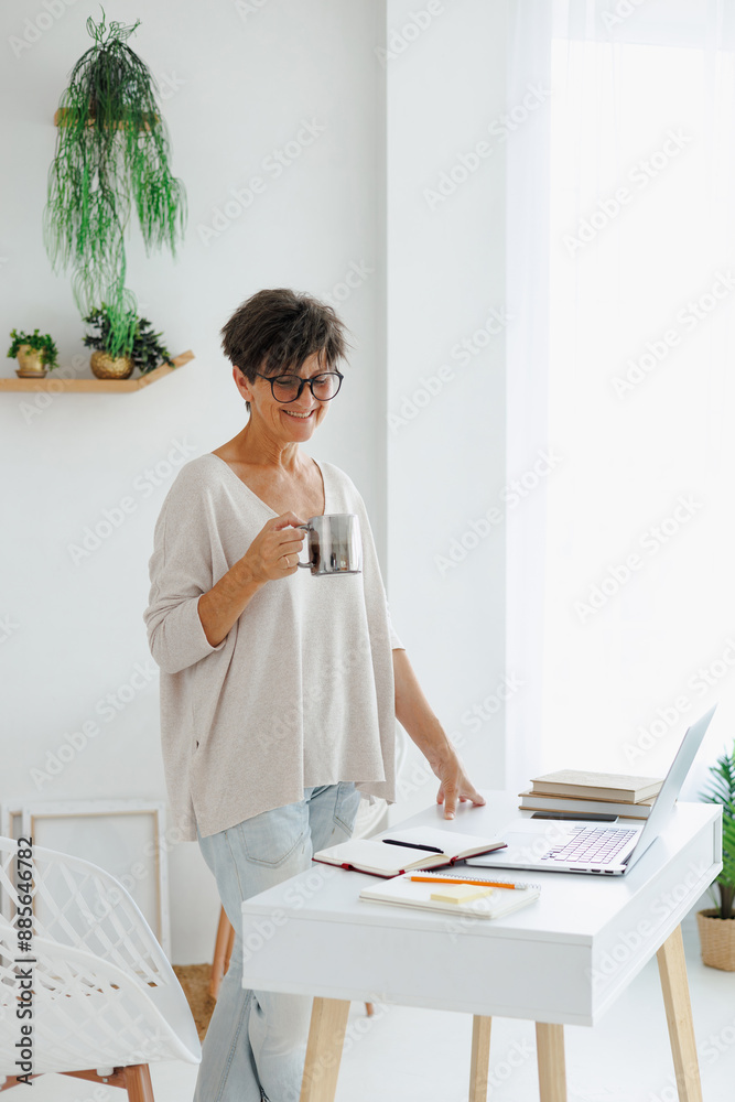 Cheerful mature woman in eyeglasses holding coffee near laptop at home Cheerful mature woman in eyeglasses holding coffee near laptop at home
