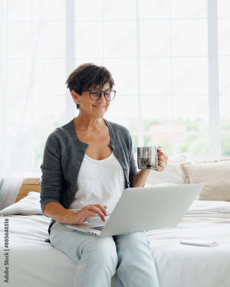 Fototapeta premium Joyful mature woman using laptop and holding coffee cup on bed at home