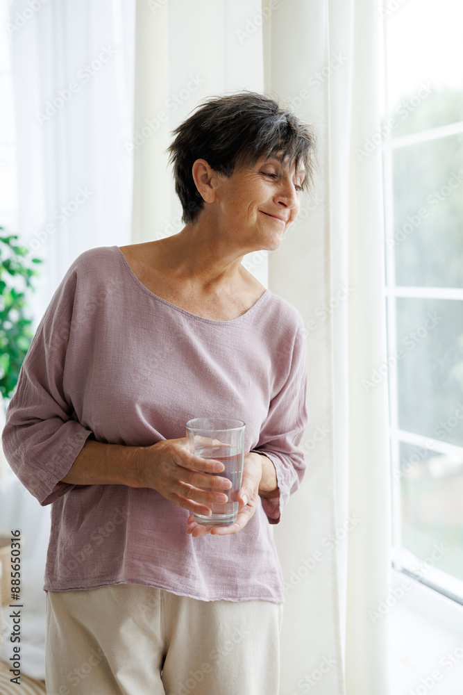 Portrait of relaxed mature woman holding glass of water near window at home Portrait of relaxed mature woman holding glass of water near window at home