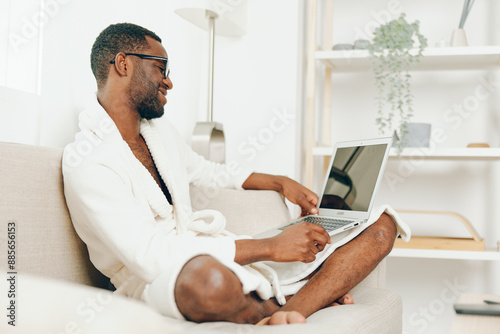 Smiling African American Freelancer Working on Laptop in Bathrobe on Sofa in the Morning The image shows a young man of African American ethnicity, dressed in a comfortable bathrobe, sitting on a © SHOTPRIME STUDIO