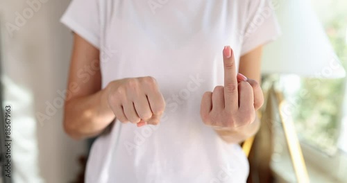 Woman in white t-shirt shows middle finger to camera standing near bright window. Female person makes obscene hand gesture slow motion