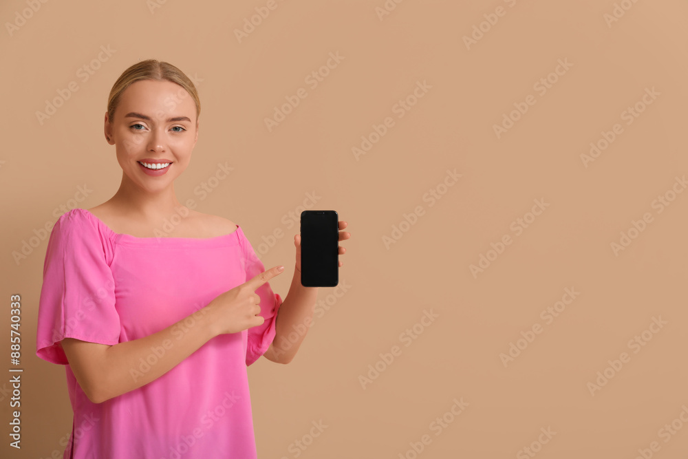 Happy young woman pointing at mobile phone with blank screen on beige background