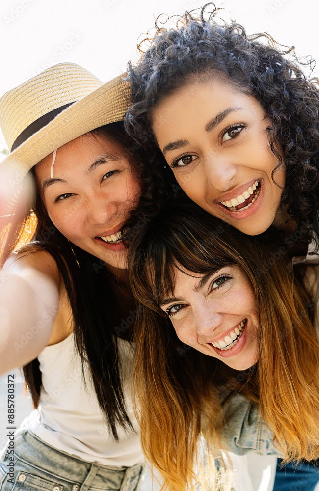 Fototapeta premium Vertical photo of three multiracial young women having fun on city street. Millennial group of female friends enjoying summer vacation taking selfie portrait outdoors. Friendship and youth concept.