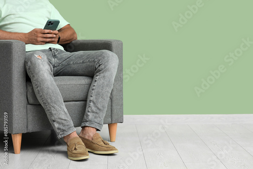 Young man in stylish shoes with mobile phone sitting on armchair near green wall, closeup