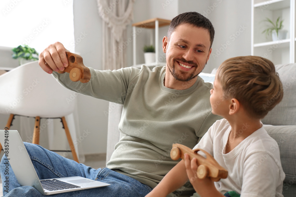 Little son and his father playing wooden toys while working with laptop at home Little son and his father playing wooden toys while working with laptop at home