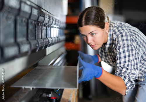 Quadro su tela Young female worker in plaid shirt and gloves operating press brake in industrial workshop, carefully positioning metal workpiece for bending on machine