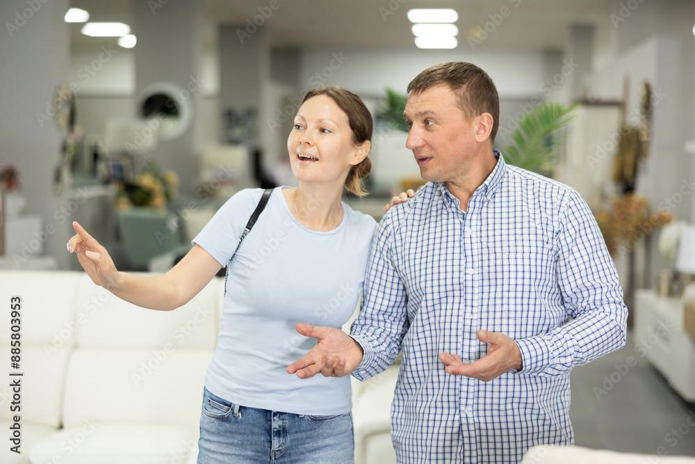 Obraz premium Spouses buyers noticed desired product in furniture store and happily smiles from upcoming purchase. Wife and husband buyer looks around in showroom and chooses right fit furniture