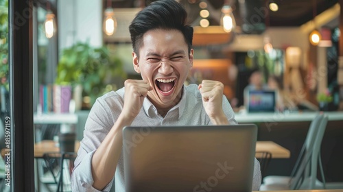 Happy young Asian businessman celebrating financial growth, staring at laptop with joy in office.