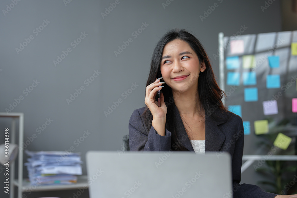 Young Asian accountant with positive attitude using smartphone at work desk. Businesswoman relaxing on mobile application talking on mobile phone and smiling while sitting in office in office. Young Asian accountant with positive attitude using smartphone at work desk. Businesswoman relaxing on mobile application talking on mobile phone and smiling while sitting in office in office.