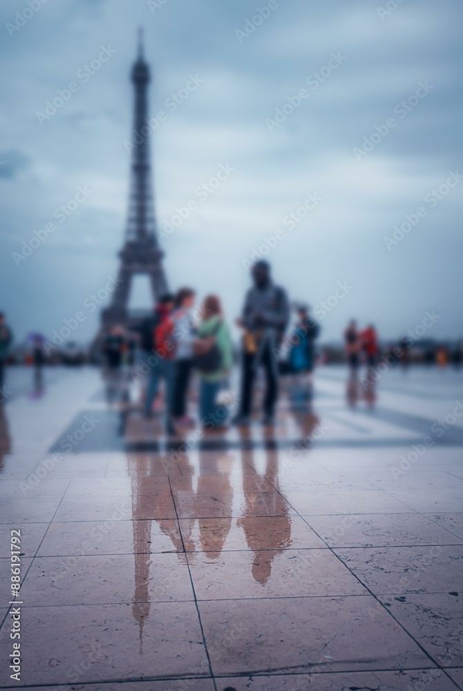 Obraz premium The Eiffel Tower is reflected in a wet Parisian square, blurred figures pass by.