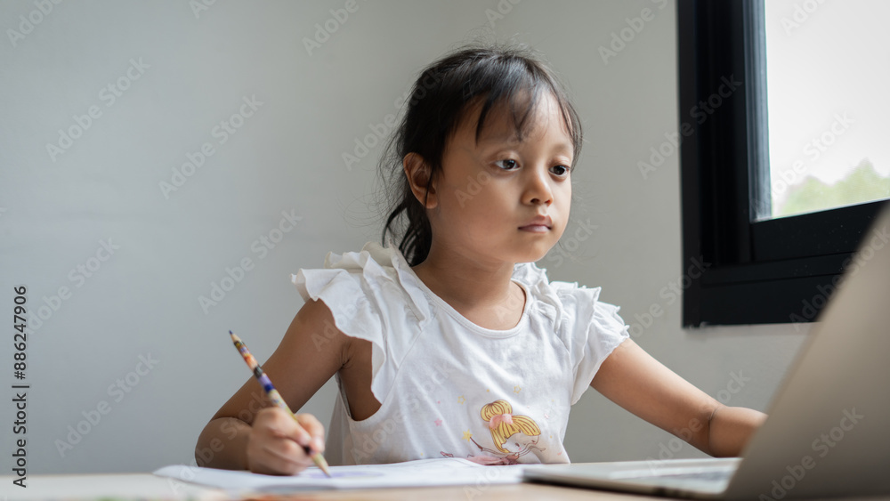 A young girl is sitting at a desk with a laptop and a pencil. She is writing on a piece of paper