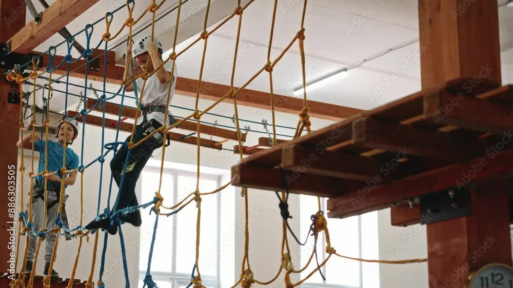 Side view from below, a boy in protective gear passes the obstacle course of the rope park. The boy overcomes an obstacle in the form of a web of ropes, purposefully and persistently. High quality 4k Side view from below, a boy in protective gear passes the obstacle course of the rope park. The boy overcomes an obstacle in the form of a web of ropes, purposefully and persistently. High quality 4k