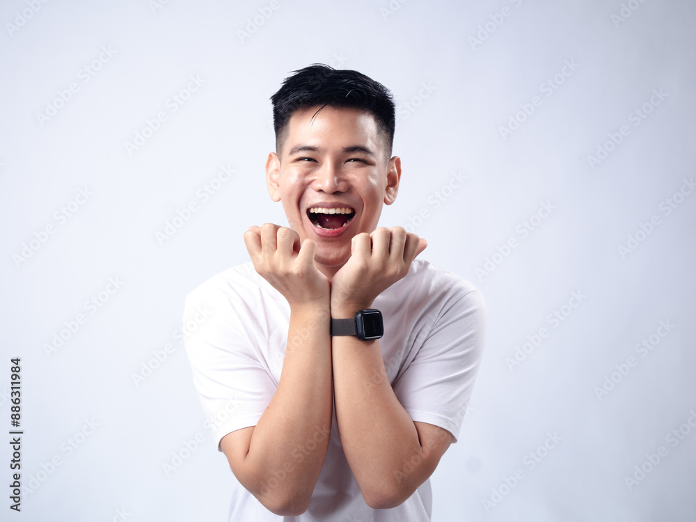 A young Asian man, wearing a white shirt and black watch, smiles broadly and holds his hands under his chin in a joyful and playful manner. The plain white background emphasizes the subject's A young Asian man, wearing a white shirt and black watch, smiles broadly and holds his hands under his chin in a joyful and playful manner. The plain white background emphasizes the subject's