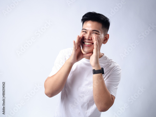 A young Asian man, wearing a white shirt and black watch, smiles widely and raises his hands near his face in an expressive and joyful manner. The plain white background highlights the subject's