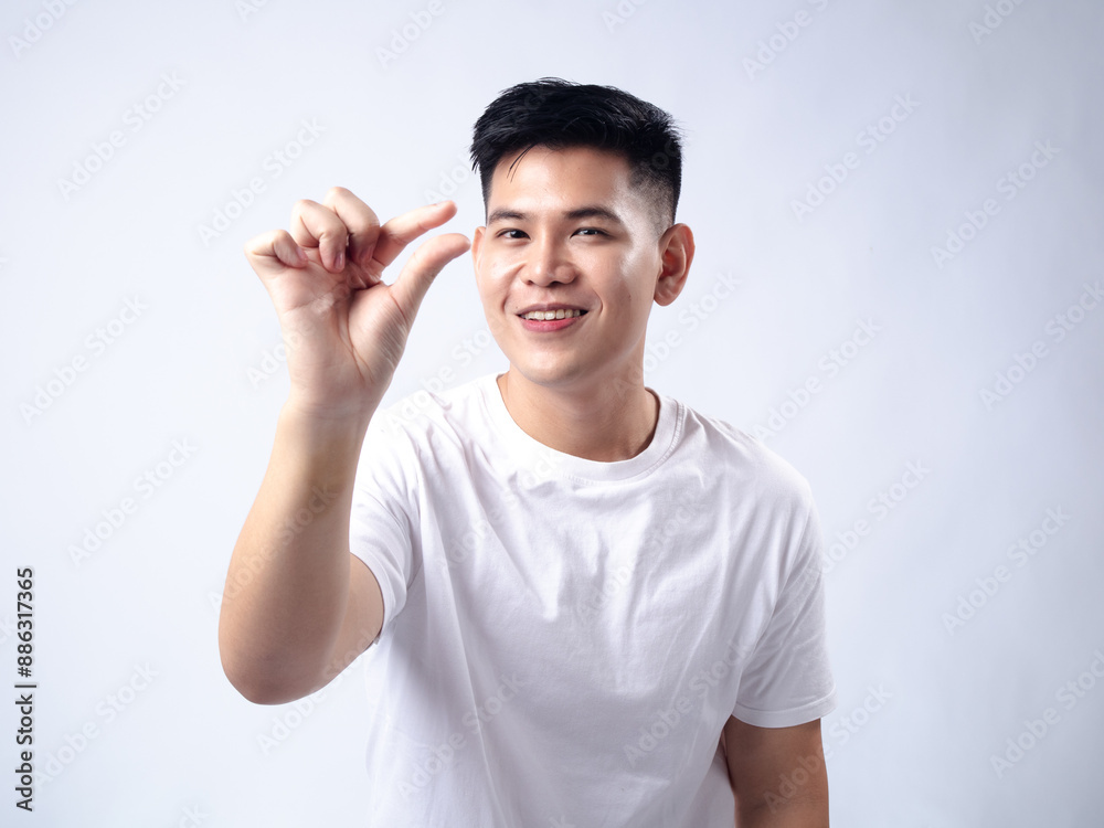 A young Asian man, wearing a white shirt, smiles while holding his fingers close together, indicating a small size. The plain white background emphasizes the subject's cheerfu A young Asian man, wearing a white shirt, smiles while holding his fingers close together, indicating a small size. The plain white background emphasizes the subject's cheerfu