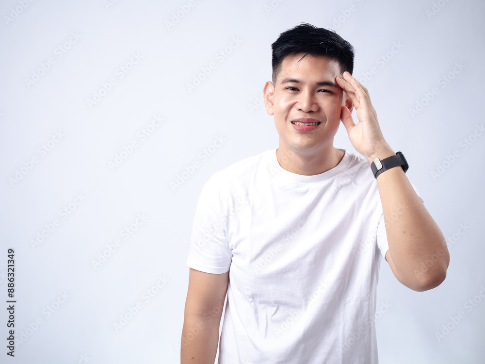 A young Asian man, wearing a white shirt and black watch, touches his temple with a slightly pained or stressed expression. The plain white background emphasizes the subject's thoughtful A young Asian man, wearing a white shirt and black watch, touches his temple with a slightly pained or stressed expression. The plain white background emphasizes the subject's thoughtful