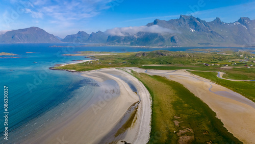 A sweeping aerial view captures a pristine sandy beach curving around a lush green coastal landscape, set against the backdrop of majestic mountains and a vast blue ocean. Kolbeinsanden Beach, Lofoten © Fokke Baarssen