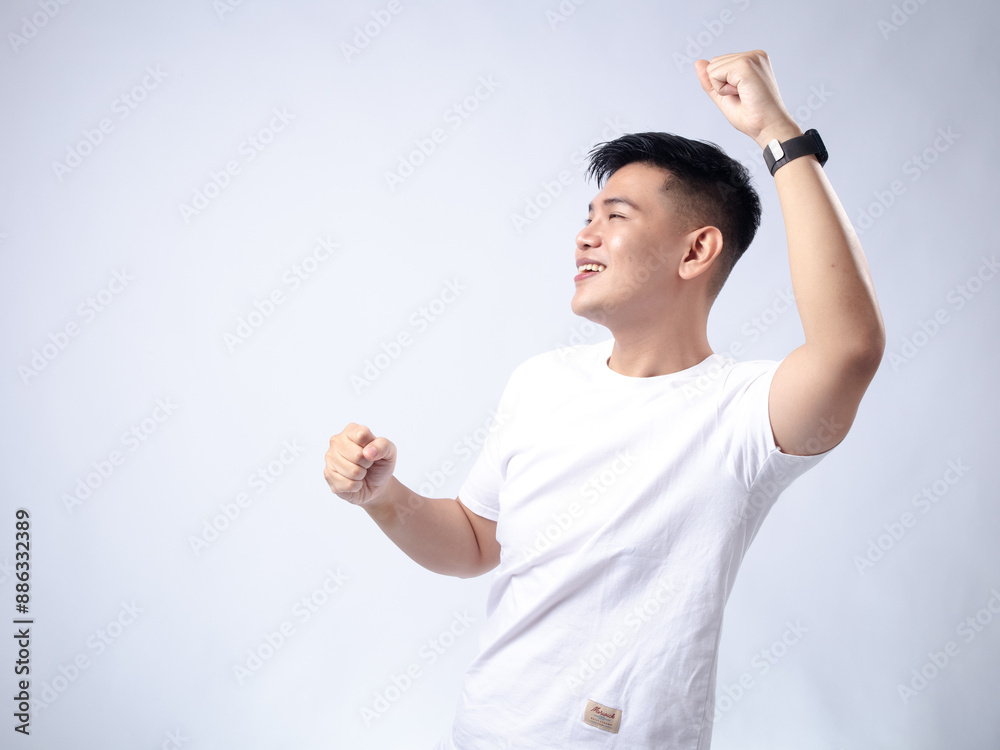A young Asian man, wearing a white shirt and black watch, smiles brightly while raising his right fist in a celebratory gesture. The background is plain white, highlighting the subject's enthusiastic A young Asian man, wearing a white shirt and black watch, smiles brightly while raising his right fist in a celebratory gesture. The background is plain white, highlighting the subject's enthusiastic