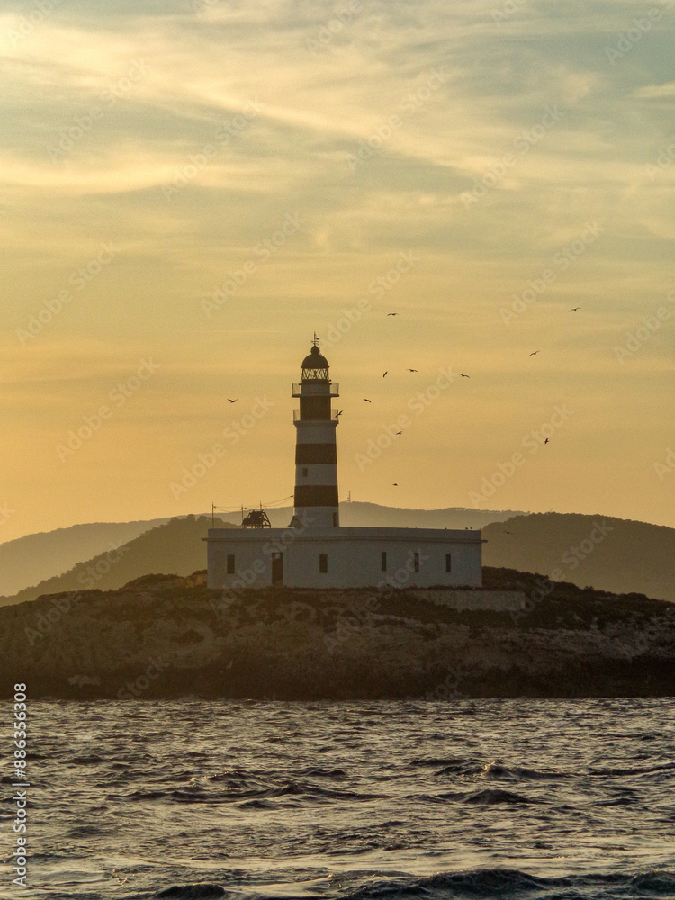 Silhouetted lighthouse on a rocky island at sunset, framed by a serene sky and gentle waves, captured off the coast of Ibiza. Silhouetted lighthouse on a rocky island at sunset, framed by a serene sky and gentle waves, captured off the coast of Ibiza.
