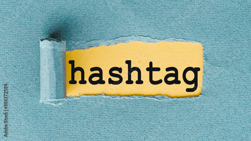 A close-up image of the word hashtag written in black text on a torn piece of yellow paper, revealing a blue background.