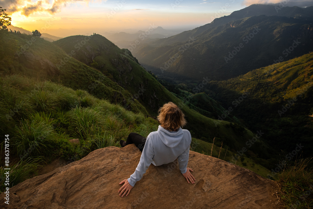Fototapeta premium A woman sits on a rock amidst the rugged terrain of the Sri Lankan mountains, taking in the panoramic view.