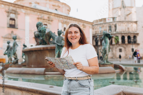 Attractive female tourist is exploring new city. Redhead happy woman holding a paper map near Fountain Rio Turia on Square of the Virgin Saint Mary, Valencia Cathedral. Traveling Europe photo