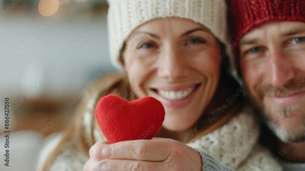 A close-up of a couple in winter hats and sweaters, smiling warmly while holding a small, red heart, radiating love, warmth, and happiness during a cozy indoor moment. A close-up of a couple in winter hats and sweaters, smiling warmly while holding a small, red heart, radiating love, warmth, and happiness during a cozy indoor moment.