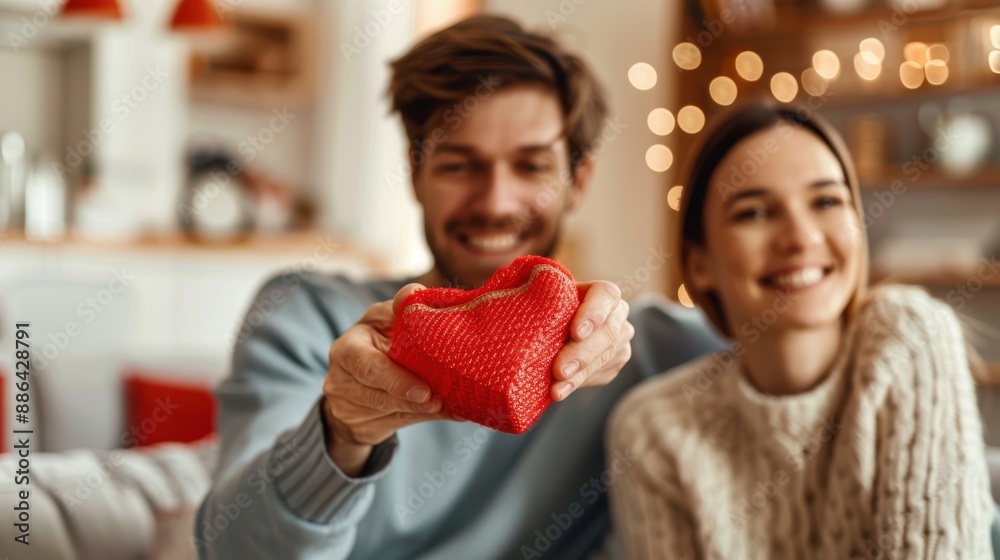 A happy couple, seated indoors, holds a small red heart-shaped gift, symbolizing love and affection in a moment of sharing and caring, illuminated by soft lights.