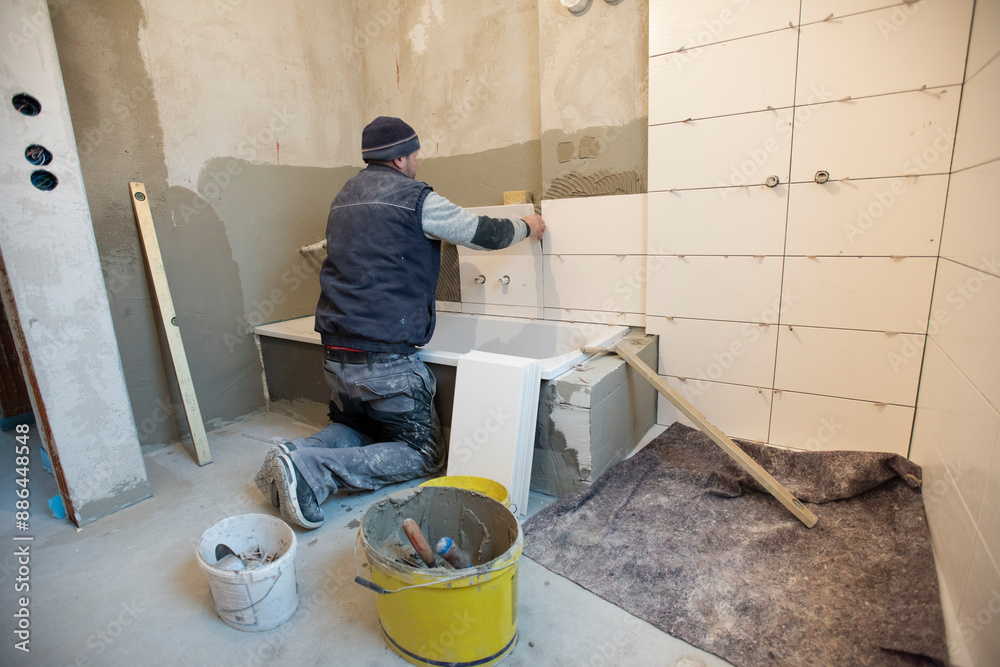 In einem Neubau klebt ein Bauarbeiter und Fliesenleger neue Fliesen an die Wand von einem Badezimmer