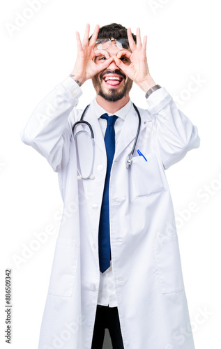 Young doctor man wearing hospital coat over isolated background doing ok gesture like binoculars sticking tongue out, eyes looking through fingers. Crazy expression.