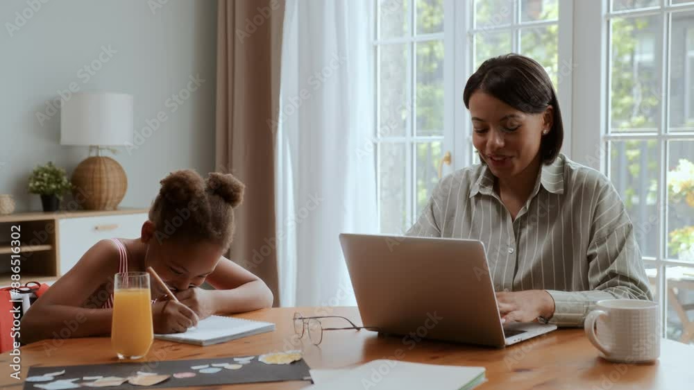 African American mother and daughter sitting at table at home. Black girl is drawing , woman is working on laptop and talks with kid smiling and enjoying, laugh together African American mother and daughter sitting at table at home. Black girl is drawing , woman is working on laptop and talks with kid smiling and enjoying, laugh together