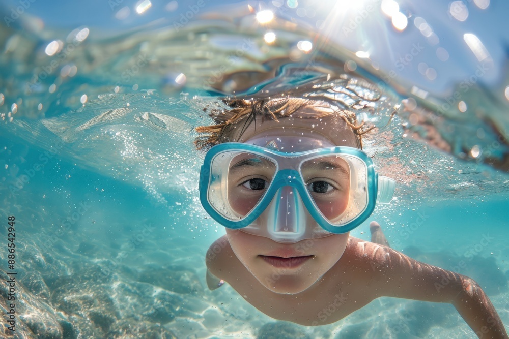 Naklejka premium A child wearing a blue snorkeling mask swims underwater in a clear turquoise sea, with sunlight creating a beautiful play of light and water around them.