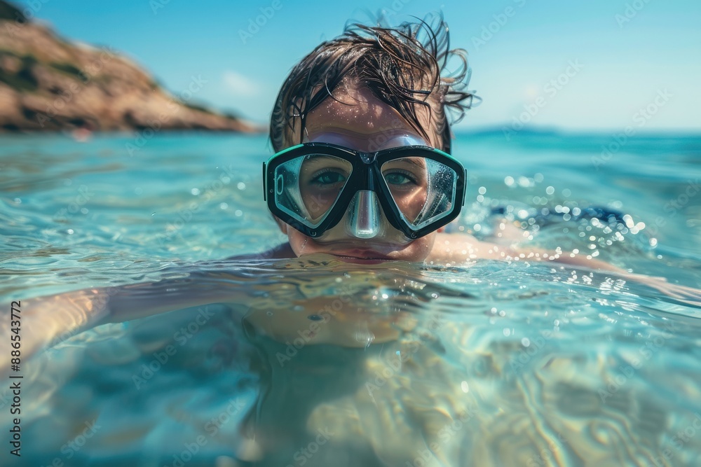 Naklejka premium A child swims in crystal clear water near the shore, wearing blue goggles, with a scenic beach in the background on a sunny day, showcasing tranquility and fun.