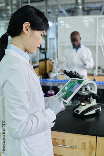 Young Chinese female scientist in gloves and lab coat pointing at screen of tablet while studying new virus or strain in laboratory