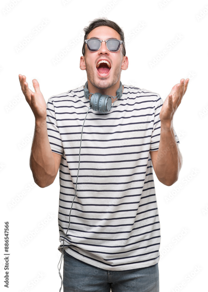 Handsome young man wearing headphones crazy and mad shouting and yelling with aggressive expression and arms raised. Frustration concept.