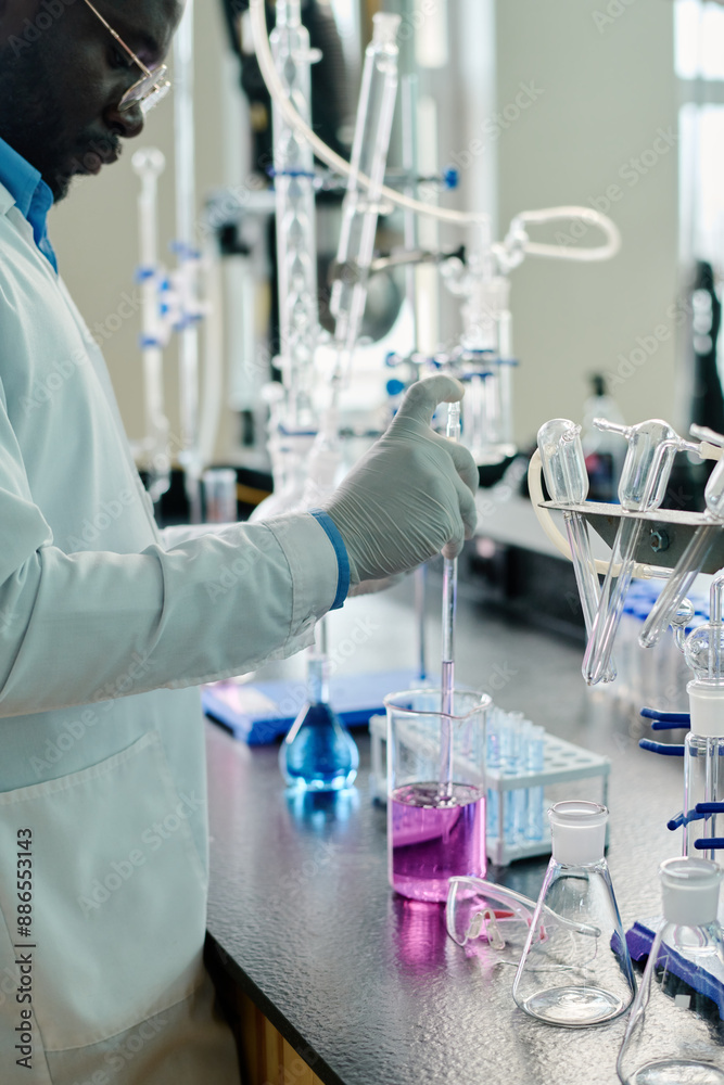 Cropped shot of gloved male scientist in lab coat standing by workplace with glassware containing liquid substances during clinical trial Cropped shot of gloved male scientist in lab coat standing by workplace with glassware containing liquid substances during clinical trial